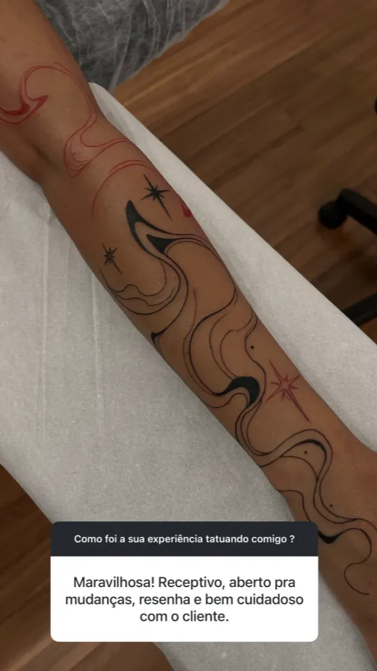 Tatuagem de linhas fluidas com estrelas, em preto e vermelho, no antebraço. Depoimento: “Maravilhosa! Receptivo, aberto pra mudanças, resenha e bem cuidadoso com o cliente.”
