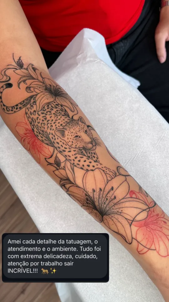 Leopardo entre lírios, em preto e vermelho, no antebraço. Depoimento: “Amei cada detalhe da tatuagem, o atendimento e o ambiente. Tudo foi com extrema delicadeza, cuidado, atenção por trabalho sair incrível!”